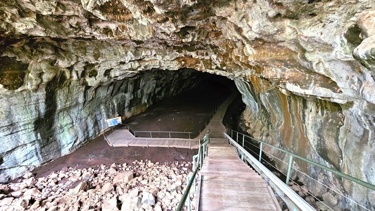 A cave interior with a rocky ceiling and walls displaying various shades of green and brown. In the foreground, a wooden staircase with metal railings descends into the cave. To the left, a fenced-off area with a sign is visible. The floor is covered with rocks and a pathway extends into the dark depths of the cave. The lighting is dim, highlighting the textures of the cave walls.