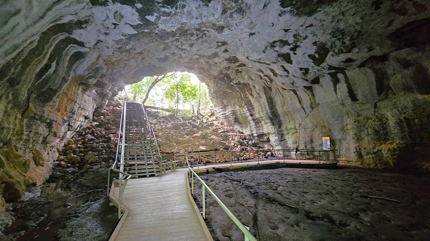A cave interior with a wooden walkway leading towards a staircase on the left. The cave walls are rugged with streaks of green and brown. The ceiling is high and uneven. At the entrance, light filters through, revealing trees and rocks outside. The floor is dark and rocky, and there is a railing along the walkway for safety.