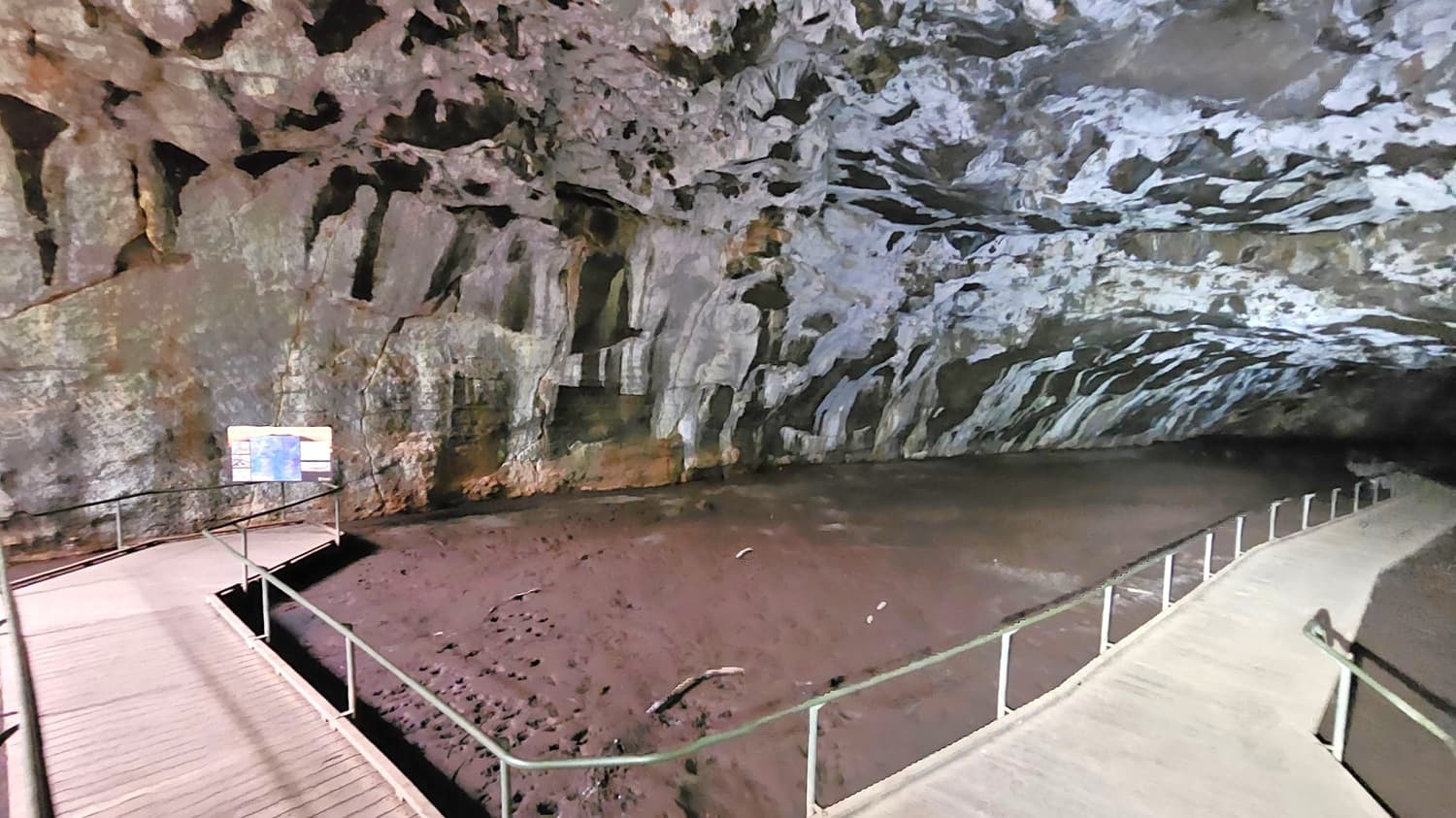 A cave interior with a wooden walkway featuring metal railings on the right side. The cave walls and ceiling are rugged, with a mix of grey, brown, and purple hues. On the left, there is an informational sign with text and images. The ground is dark and uneven, with some scattered debris. The lighting is dim, highlighting the textures of the cave surfaces.