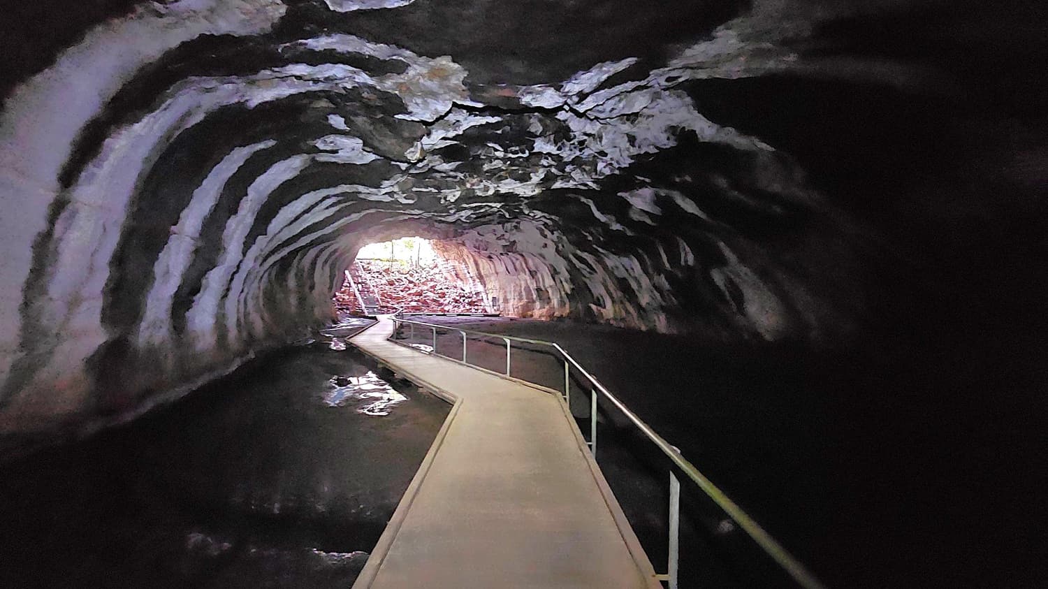 A tunnel with a curved, textured ceiling and walls, featuring a narrow walkway with metal railings running through the centre. The floor is dark, possibly water-covered. At the far end, natural light illuminates the entrance, revealing rocky terrain outside. The tunnel's interior is mostly in shadow, with the light creating a contrast at the exit.
