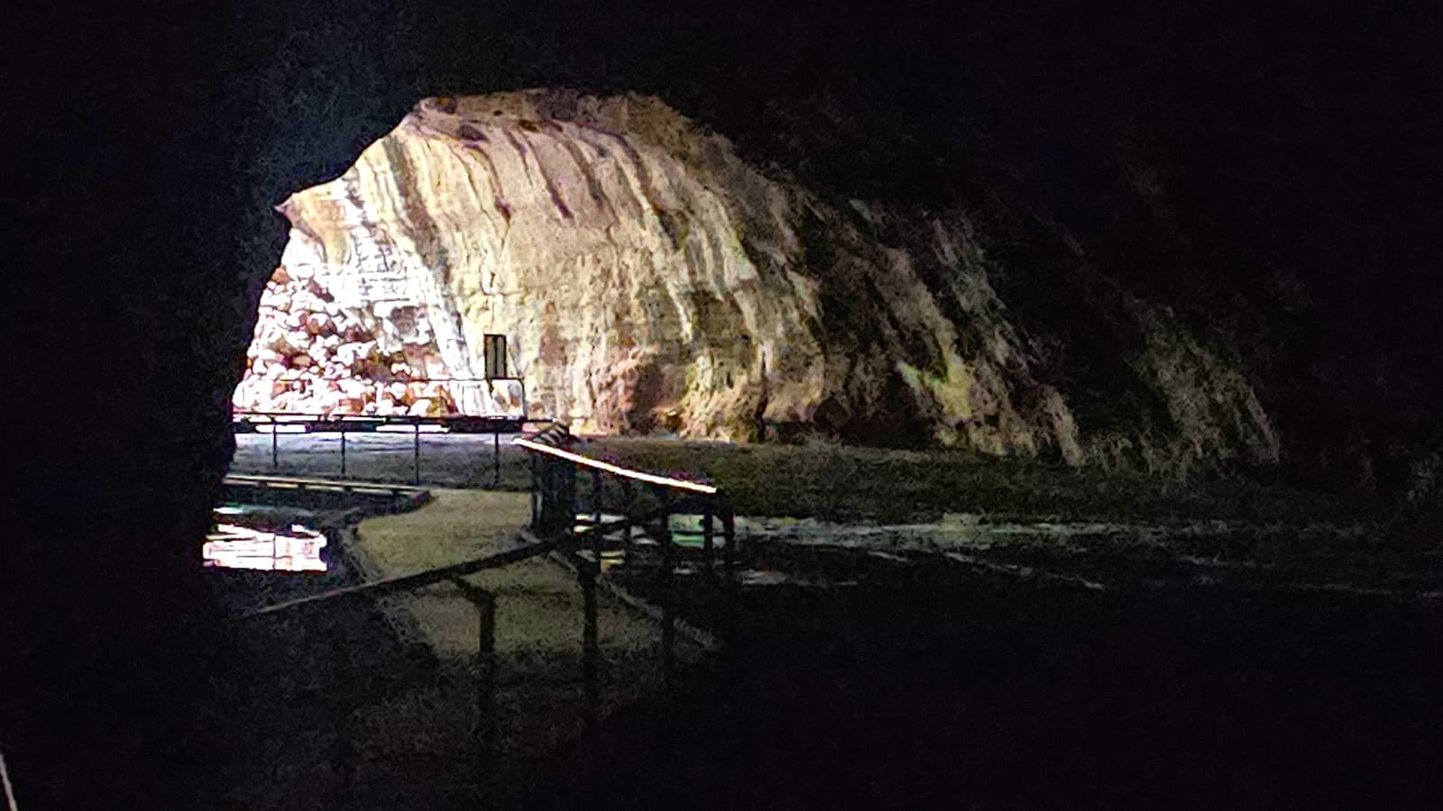 A dimly lit cave interior with a rocky wall on the right, illuminated by light from the entrance. The left side is mostly in shadow. A pathway with railings runs through the cave, leading towards the entrance. The floor appears uneven, with some reflections suggesting water presence. The ceiling is not visible due to darkness.