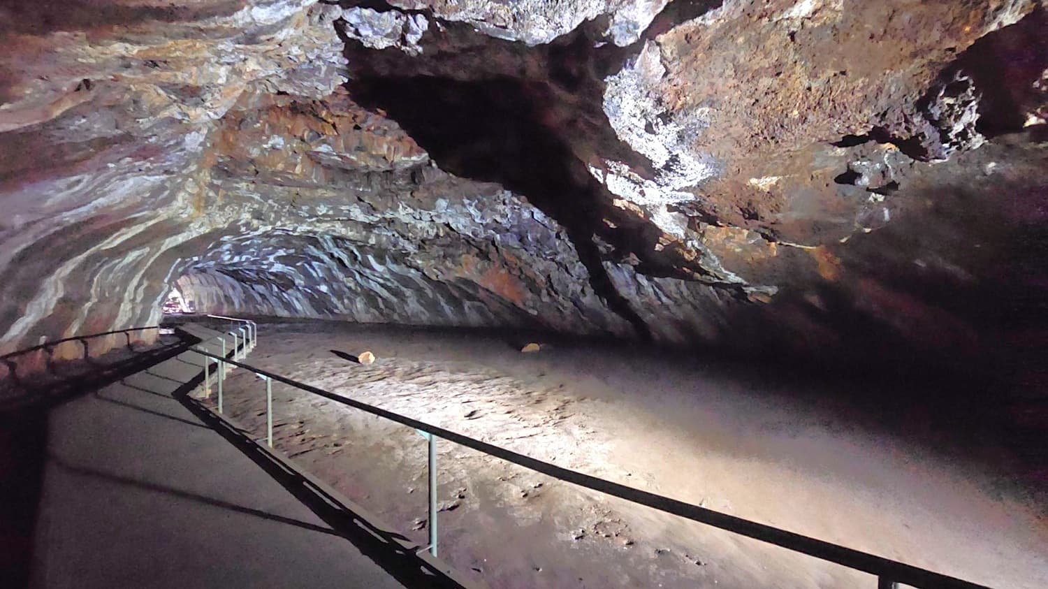 A cave interior with a rough, textured ceiling and walls displaying various shades of brown and grey. On the left, a paved walkway with a metal railing curves into the distance. The floor is uneven and sandy, with a few scattered rocks. The lighting highlights the natural formations, creating shadows and depth.
