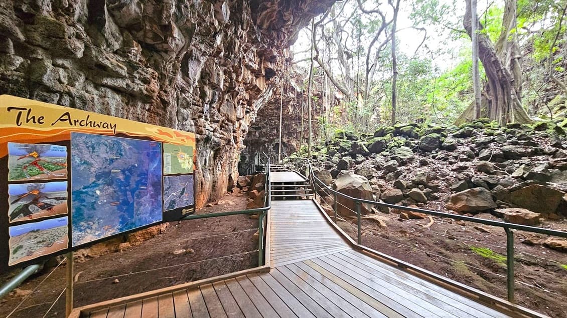 A wooden walkway with railings leads through a rocky cave area. On the left, a large informational sign titled "The Archway" displays maps and images. The cave wall is rugged and textured. To the right, there are scattered rocks and lush green trees, with sunlight filtering through the foliage. The path ascends with steps further into the cave, surrounded by natural rock formations and vegetation.