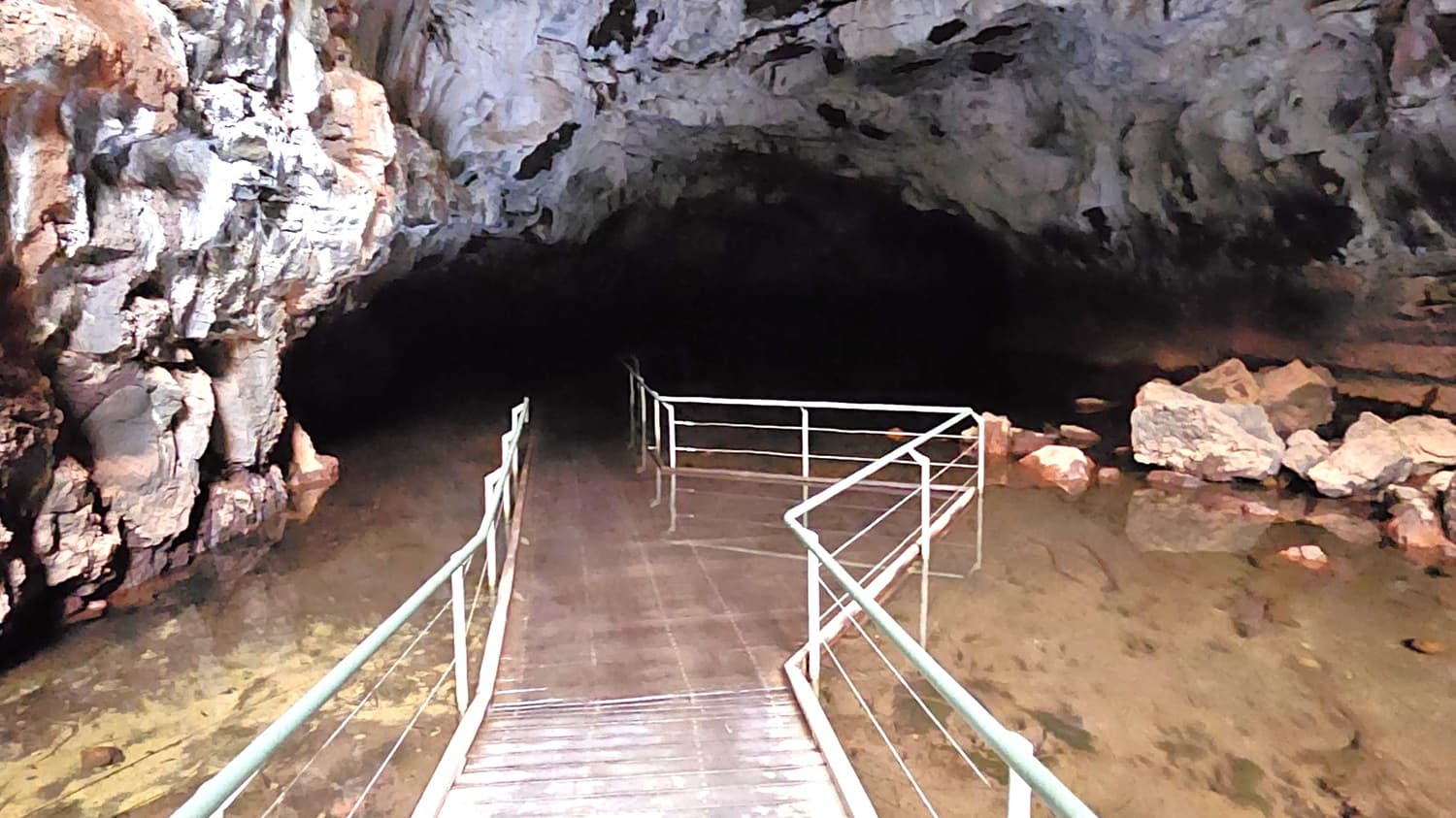A cave interior with a wooden walkway leading into the darkness. The walkway has metal railings on both sides and is partially submerged in clear water. On the left, rugged rock formations rise from the water, while on the right, there are scattered large rocks and boulders. The cave ceiling is high and uneven, with dark patches and a rough texture. The overall atmosphere is dimly lit, with natural rock colours dominating the scene.
