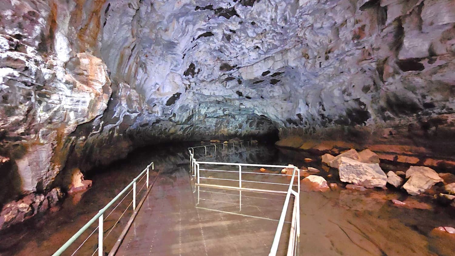 Inside a cave with rugged, textured walls in shades of grey and brown. The ceiling is high and uneven, with dark patches. A narrow metal walkway with railings extends into the cave, partially submerged in water. Large rocks are scattered on the right side, partially submerged. The lighting is dim, casting shadows and highlighting the cave's natural formations.