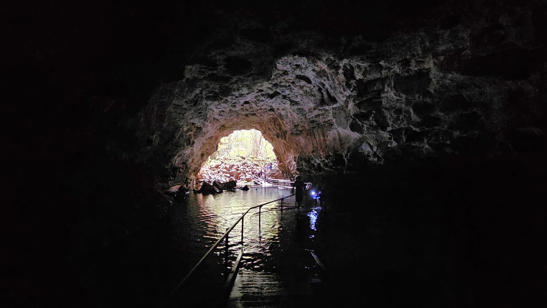 A dark cave interior with a rocky ceiling and walls. In the centre, a bright opening reveals the outside, with rocks and vegetation visible. Water covers the cave floor, reflecting the light from the entrance. A metal handrail runs along a submerged path leading towards the exit. A person with a light is visible near the entrance, adding a sense of scale to the scene.