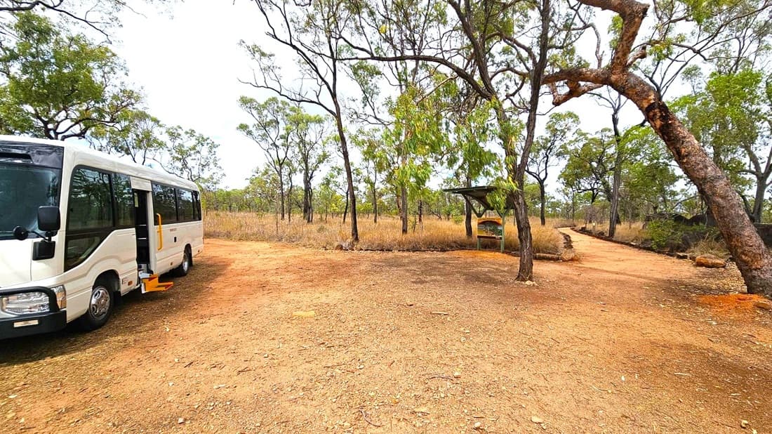 A white bus is parked on the left with its door open and a yellow step extended. The ground is covered in reddish-brown dirt. In the centre, there are scattered trees with sparse foliage. On the right, a dirt path leads into the distance, flanked by more trees and dry grass. A small shelter with a sign is visible near the path. The scene is set in a natural, wooded area.