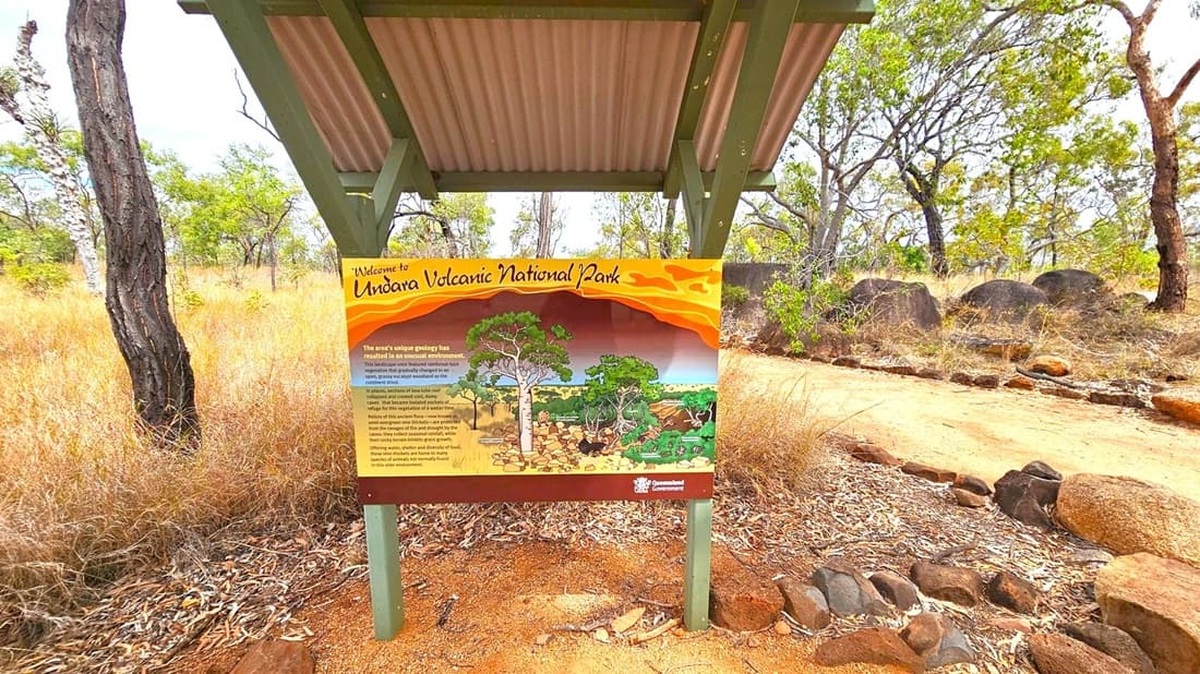 A signboard under a corrugated metal shelter welcomes visitors to Undara Volcanic National Park. The sign features an illustration of the park's landscape and information about its unique geology. Surrounding the sign are dry grasses and scattered rocks. In the background, there are trees with sparse foliage, and a dirt path runs alongside the sign. The ground is covered with dry leaves and small stones.