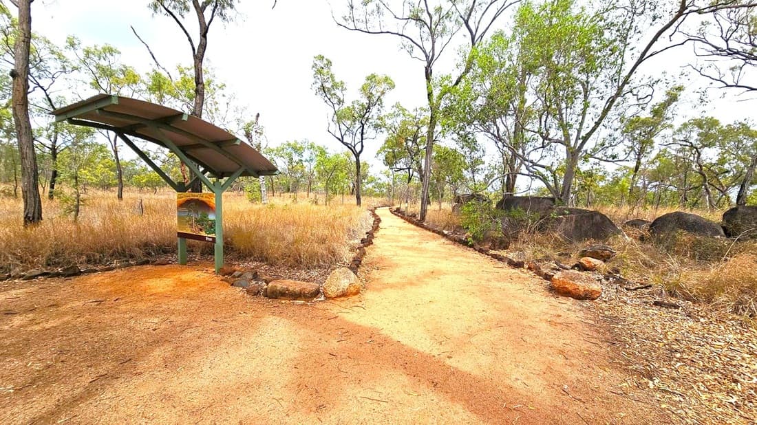 A dirt path winds through a dry, grassy landscape with scattered trees. On the left, a covered information board stands, providing details about the area. The path is bordered by rocks and leads into the distance, flanked by more trees and large boulders on the right. The ground is a mix of reddish and brown earth, with dry leaves scattered around. The sky is overcast, casting a muted light over the scene.