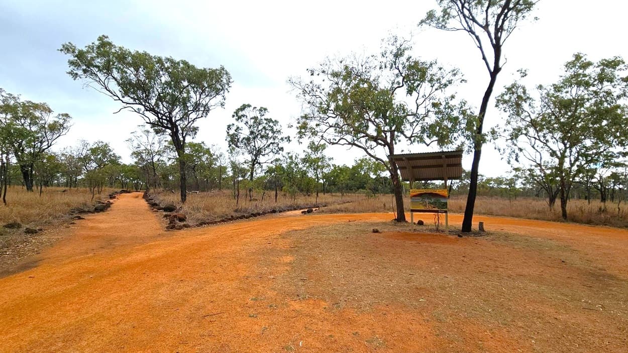 A dirt path curves through a dry, grassy landscape with scattered trees. On the right, a small shelter with a corrugated metal roof stands beside a tree, with a sign beneath it. The ground is a mix of reddish and brown hues, with patches of green. The background is filled with more trees and dry grass, creating a sparse, open environment.