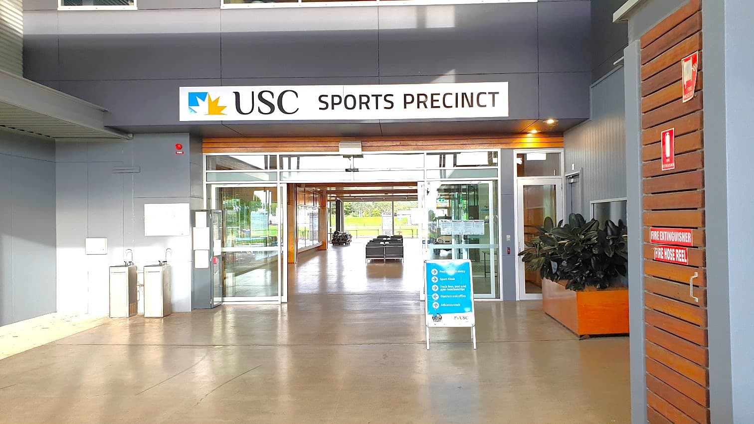 The main entrance with 'USC sports precinct' written up and an automated glass door is located on the pathway to a well-lit space with a row of seats in the centre, leading to a field at the end. To the left, 2 water fountains are located, whilst on the right there is a a notice board, signage for a fire alarm, and an infographic on the wall.