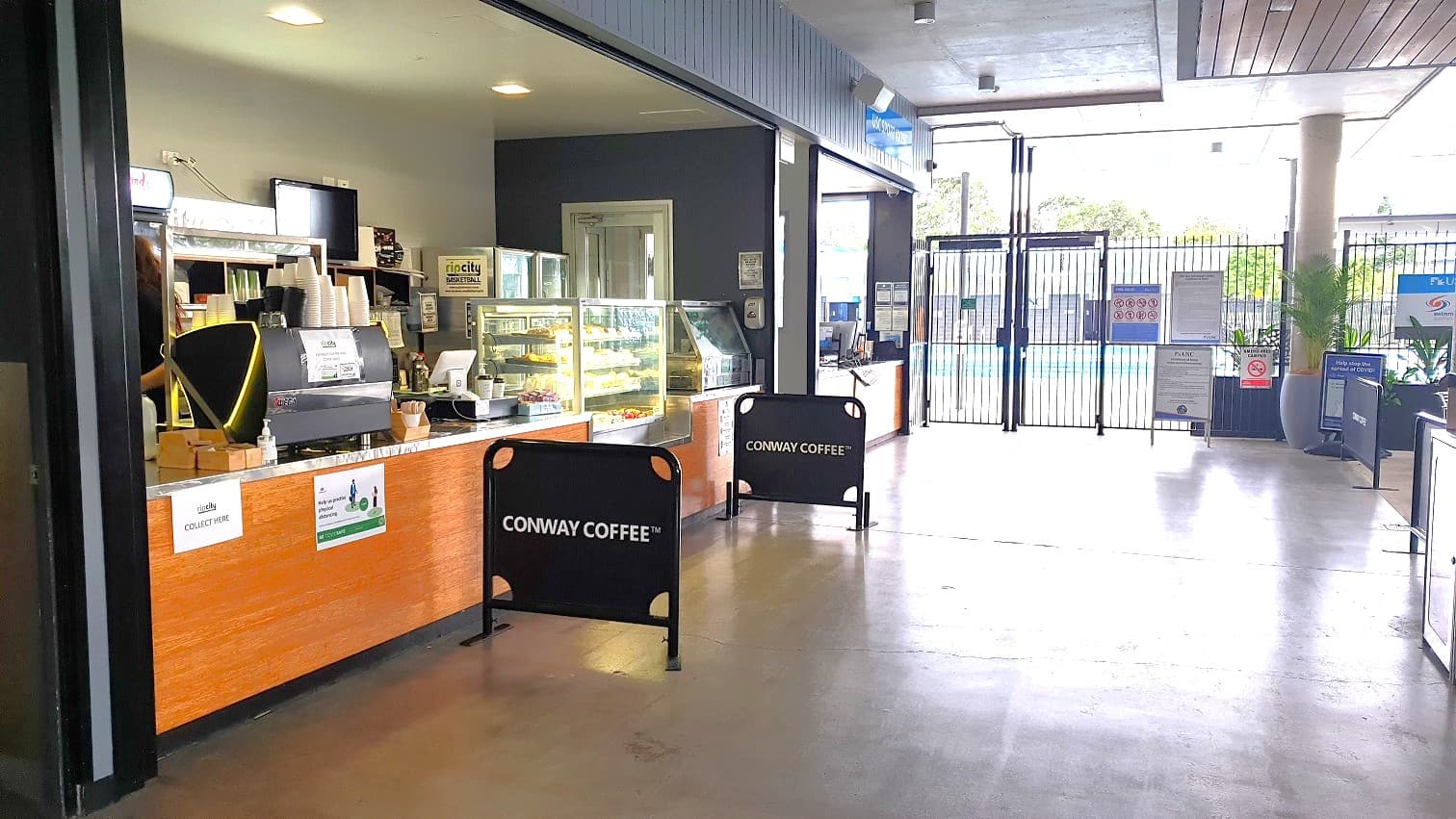 To the left of the see-through fence leading to the pool is a retail area with a café counter and another cubicle with infographics on the wall. The café has a TV on the wall, a reception counter stocked with snacks in a glass cabinet, an ice cream machine and cups, a desktop, and a few boxes. There are Conway Coffee signage at near the counter for customers to queue alongside. A turnstile gate is to the right, with a large palm in a grey pot beside the pillar with signage on the wall.
