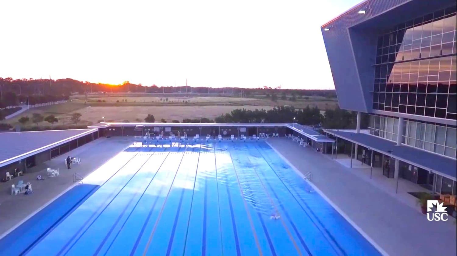 A 10-lane open pool with a four-story grey structure to the right that has entrances going to the pool area. To the left is a covered sitting area with white chairs and a table, and to the right is an outdoor sitting area with white chairs and a table.