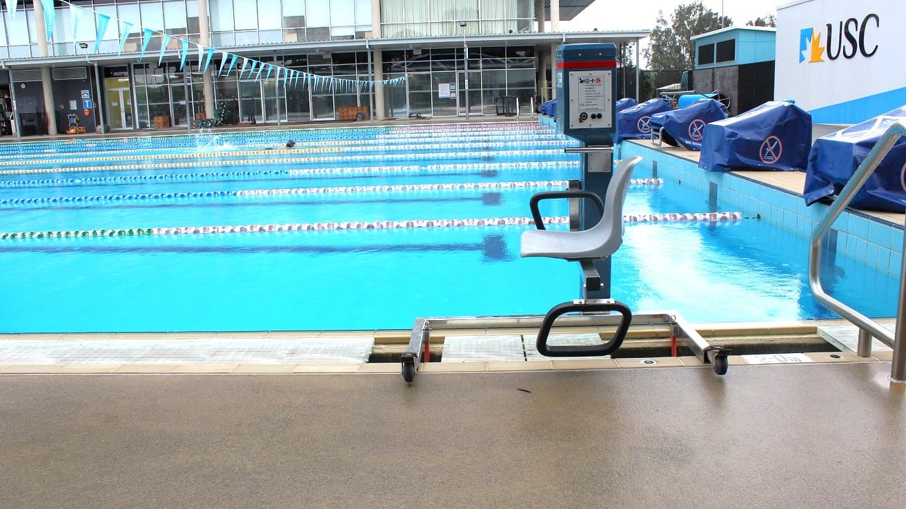 A large, blue open swimming pool with blue, pink, and green ropes separating the lanes with a line of small, blue flags hanging across the pool. There is a hoist positioned directly next to the pool. To the right is a USC branded wall with the swimming blocks covered in blue bags with an icon representing 'no diving'. The pool looks into a grey glass structure with entrance doors.