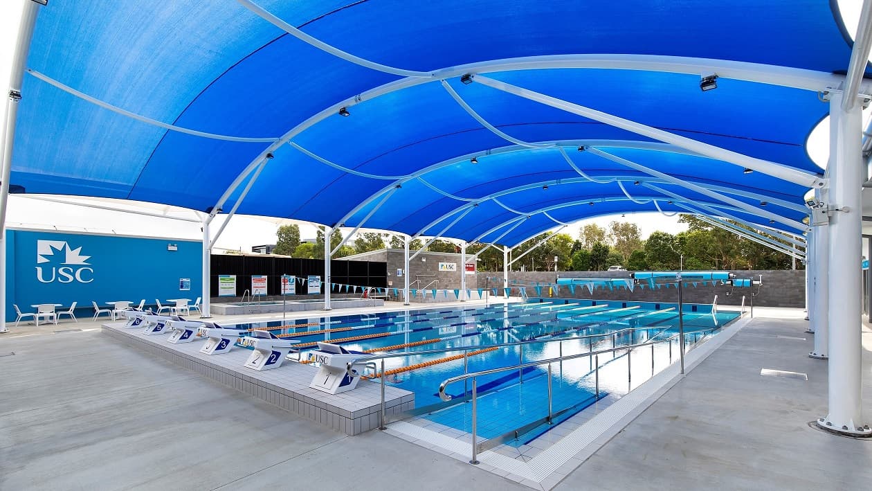 A six-lane pool in a blue tunnel-like shade that is lit with natural light. There are white and light blue flags hanging across the pool. To the left is a sitting area with seats and a table, with the USC symbol painted on the blue wall. There are black gloss walls with signage on them. The brick structure towards the end has the USC and a muster point sign. The area is enclosed by a brick wall.