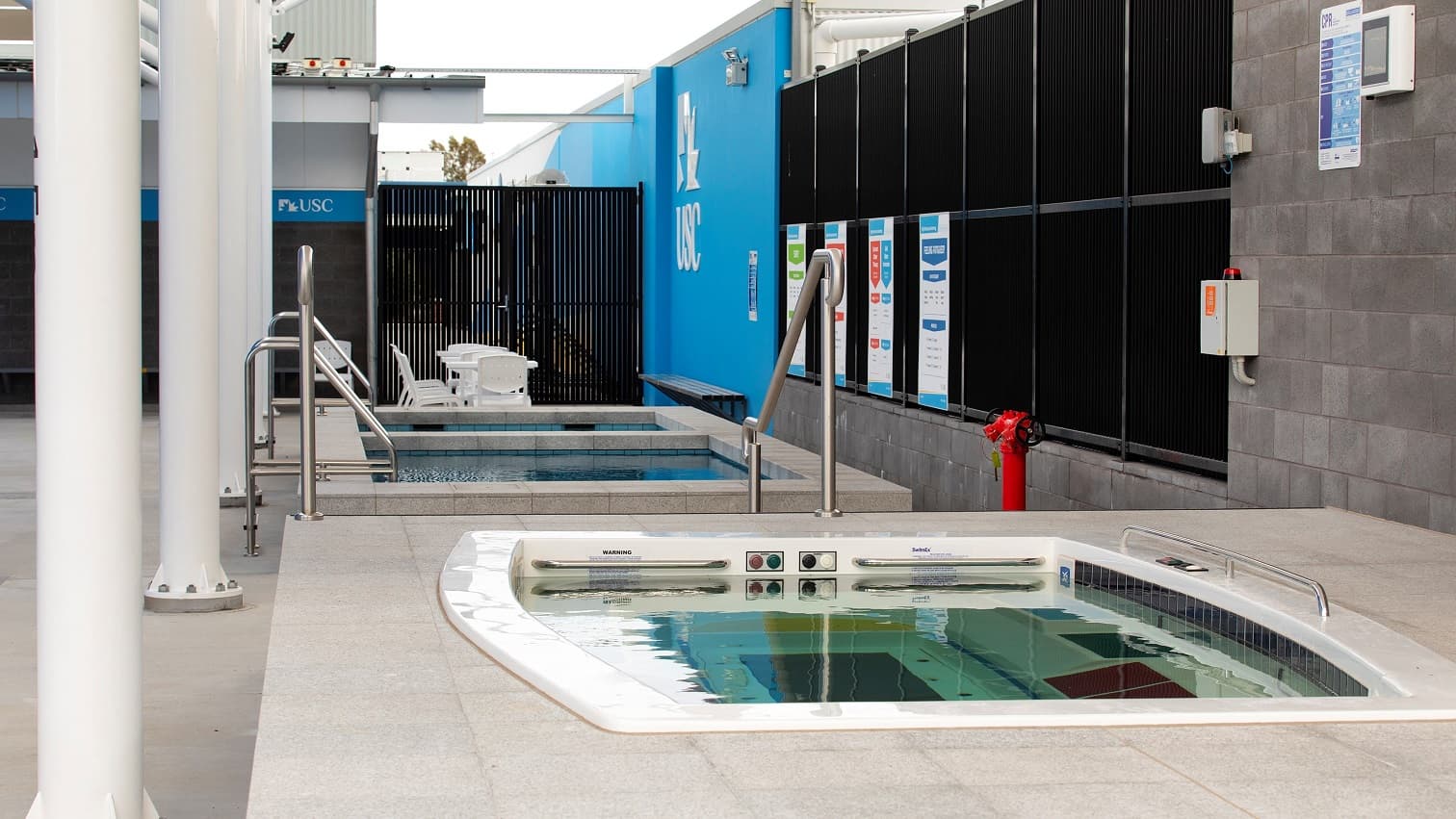 In an open area, is a hydrotherapy pool facing a cool and a warm plunge pool. Towards the end of the space, there is a seating area with white plastic chairs and a plastic table facing a black fence. On the left side, there is a table leaning against the blue wall. The right side has a black glass wall with signage and a fire hydrant. There is an emergency alarm on the brick wall.