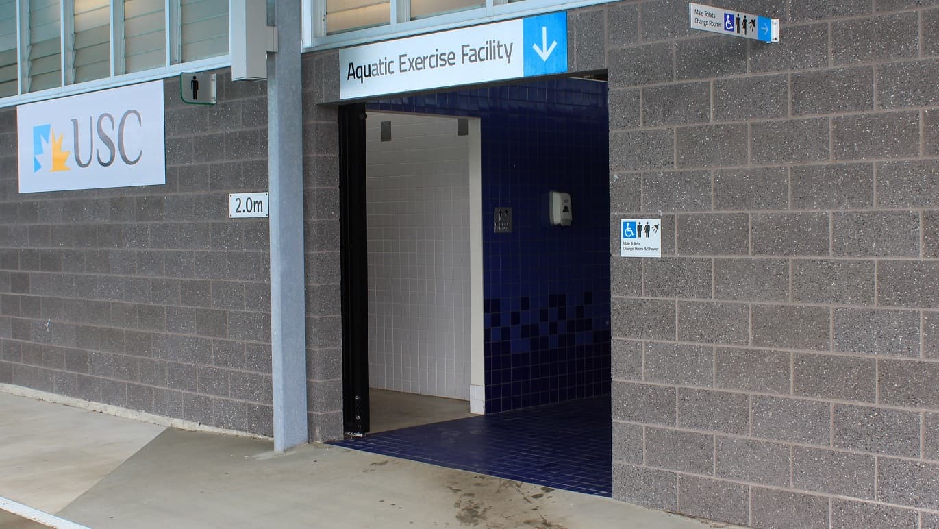 A blue-tiled entrance with a sign above stating 'Aquatic Exercise Facility' and an arrow pointing down. A white tiled entryway leads to the male restroom and change room on the left. A sign detailing 'USC' is on the left.