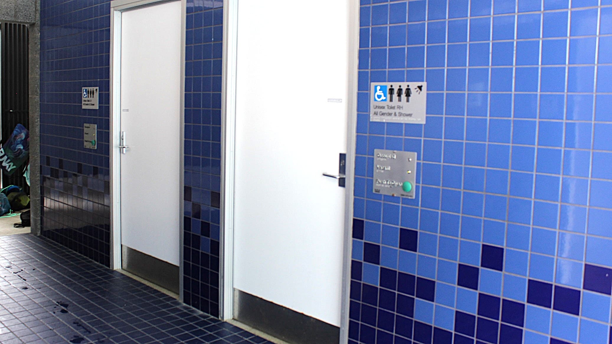 This area features unisex toilets and shower rooms. The floor and walls are adorned with dark blue and light blue tiles, while the doors are white in color. The wall displays the toilet signage to the side of each of the two doors.