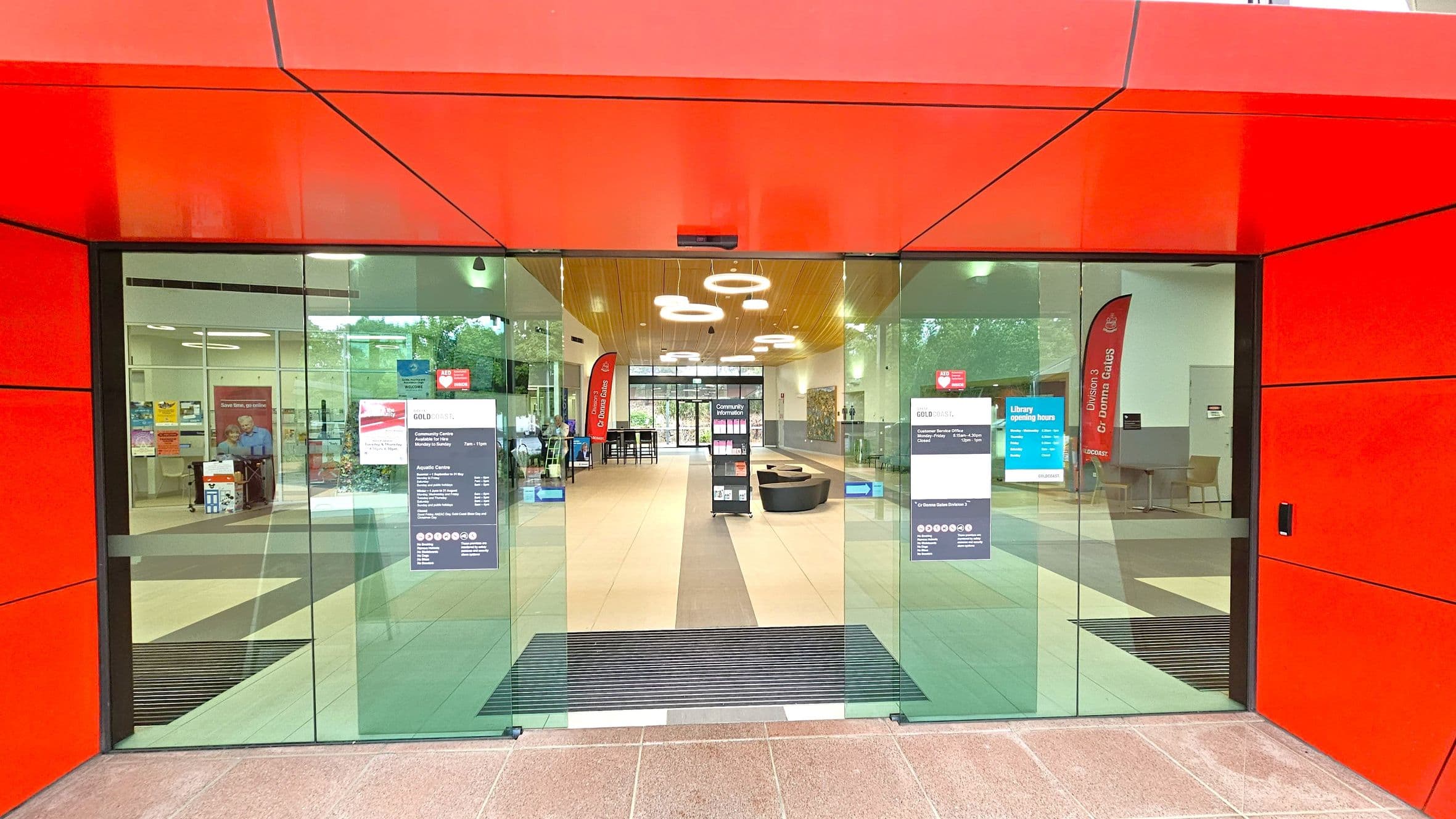 A modern entrance with large glass doors framed by bright red panels. Inside, the floor is tiled, and the ceiling features circular light fixtures. On the left, there are posters and a seating area with a person visible. On the right, a red vertical banner and more seating are present. In the centre, a display stand and a pathway lead to another set of glass doors at the back, revealing greenery outside.