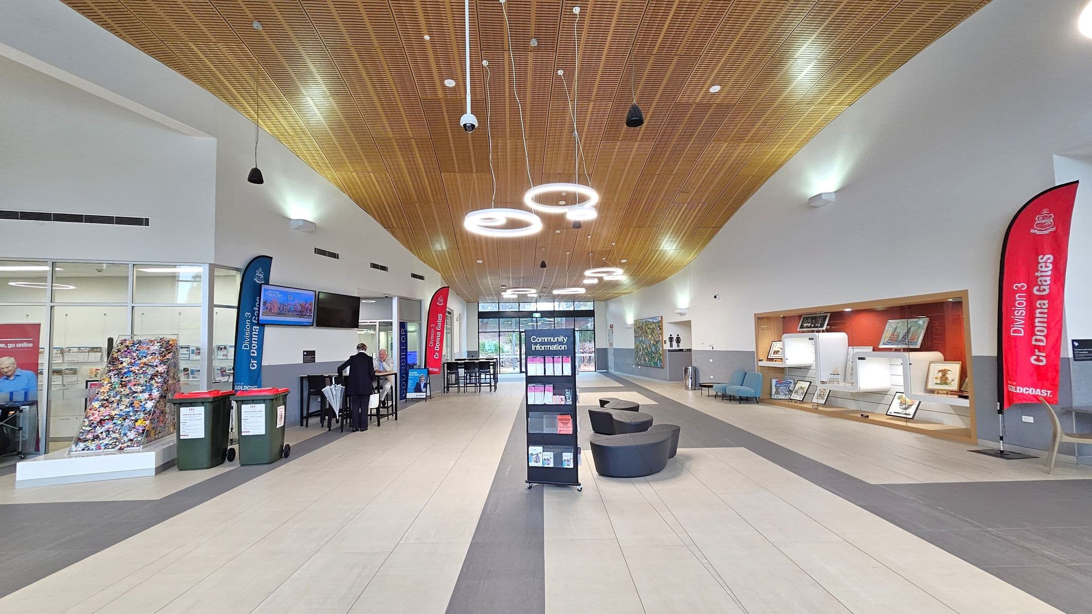 A spacious indoor area with a wooden panelled ceiling featuring circular lights. In the centre, a stand labelled "Community Information" holds brochures. On the left, a glass wall with posters and two large bins. A person is seated at a table. On the right, a display case with artwork and a red banner. The floor is tiled in light and dark grey. At the back, glass doors lead outside, flanked by more seating and artwork.