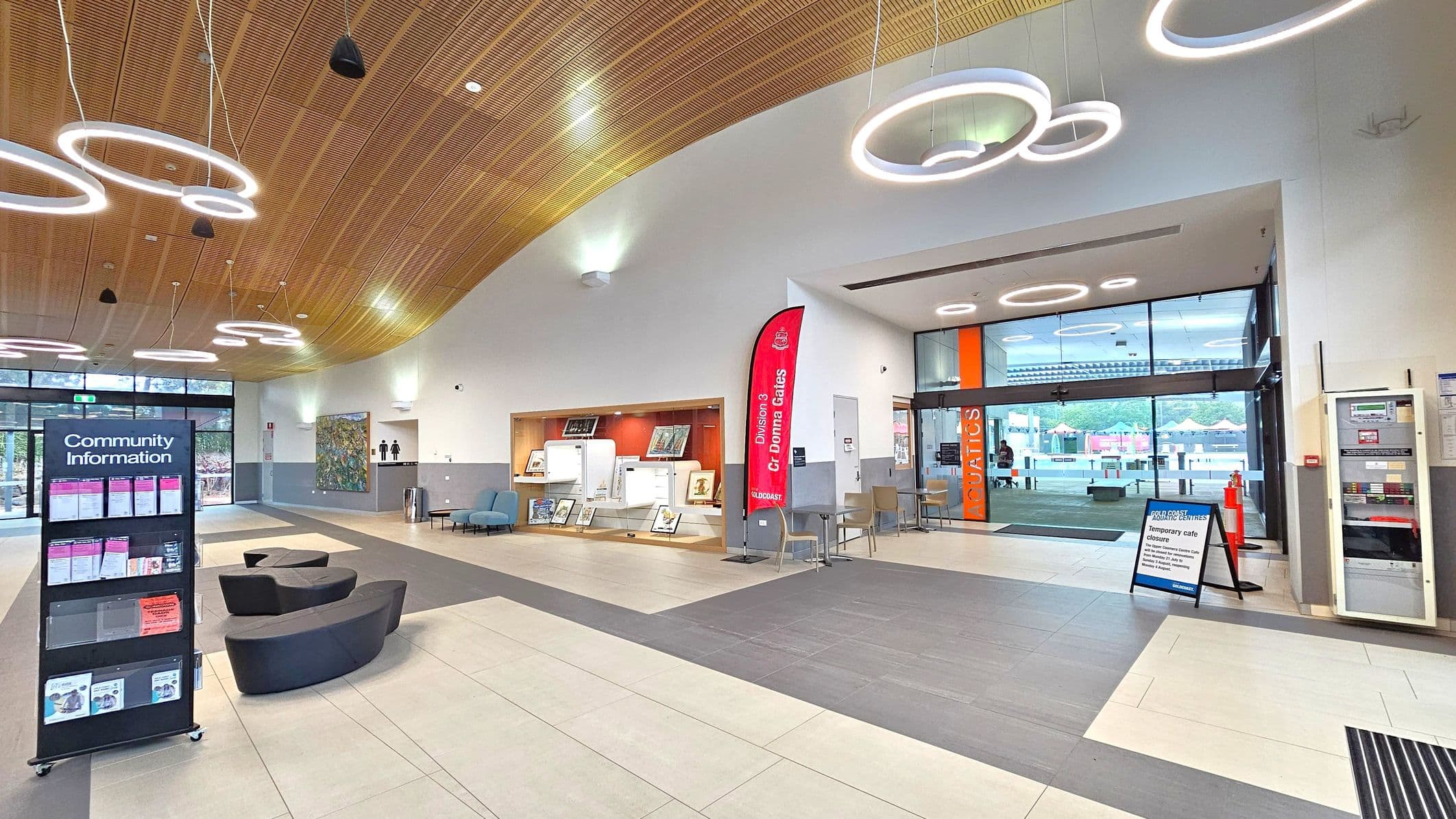 A modern interior with a curved wooden ceiling featuring circular light fixtures. On the left, a community information stand with brochures and curved black seating. In the centre, a display area with framed items and a red banner. On the right, glass doors with "AQUATICS" signage lead outside, with a temporary café closure sign nearby. The floor is tiled in beige and grey, and there are chairs and a vending machine near the entrance.