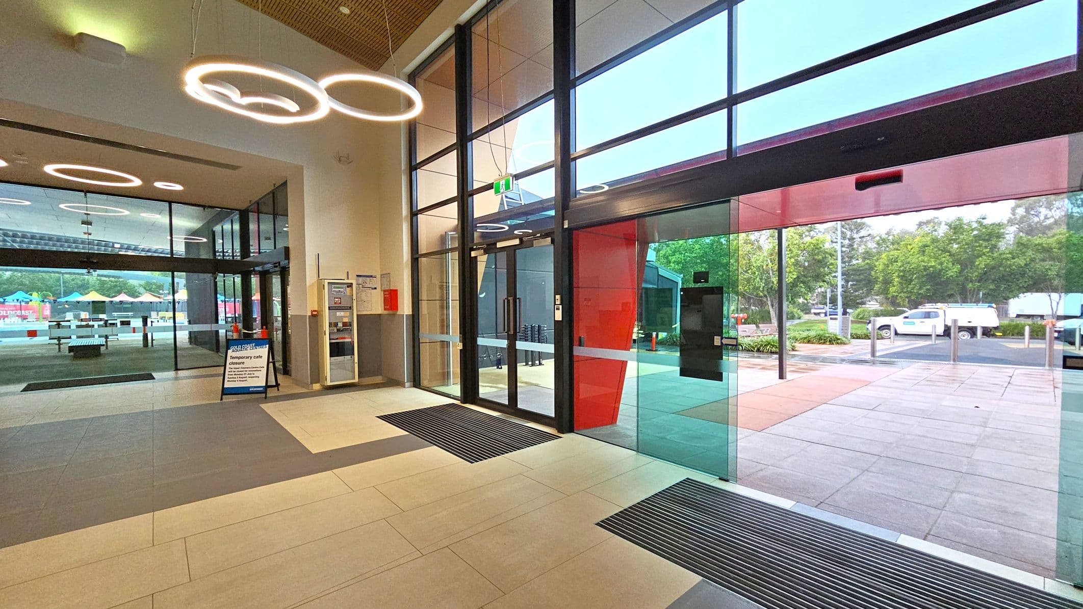 A modern interior with large glass doors on the right leading outside, where trees and a parked vehicle are visible. The floor is tiled in light and dark grey. On the left, a glass wall separates another area with colourful umbrellas and tables. Circular ceiling lights hang above, and a sign on the floor indicates a temporary café closure. A vending machine and fire safety equipment are mounted on the wall near the entrance.