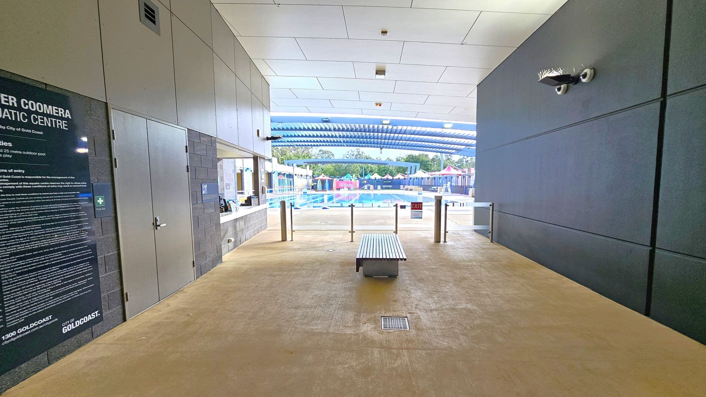 A corridor leading to an outdoor swimming pool. On the left, there's a large sign with text and a door. On the right, a dark wall with a security camera. In the centre, a bench is placed on a light-coloured floor. Beyond the corridor, the pool area is visible with a blue and white striped canopy, colourful structures, and trees in the background. The ceiling is white with recessed lighting.