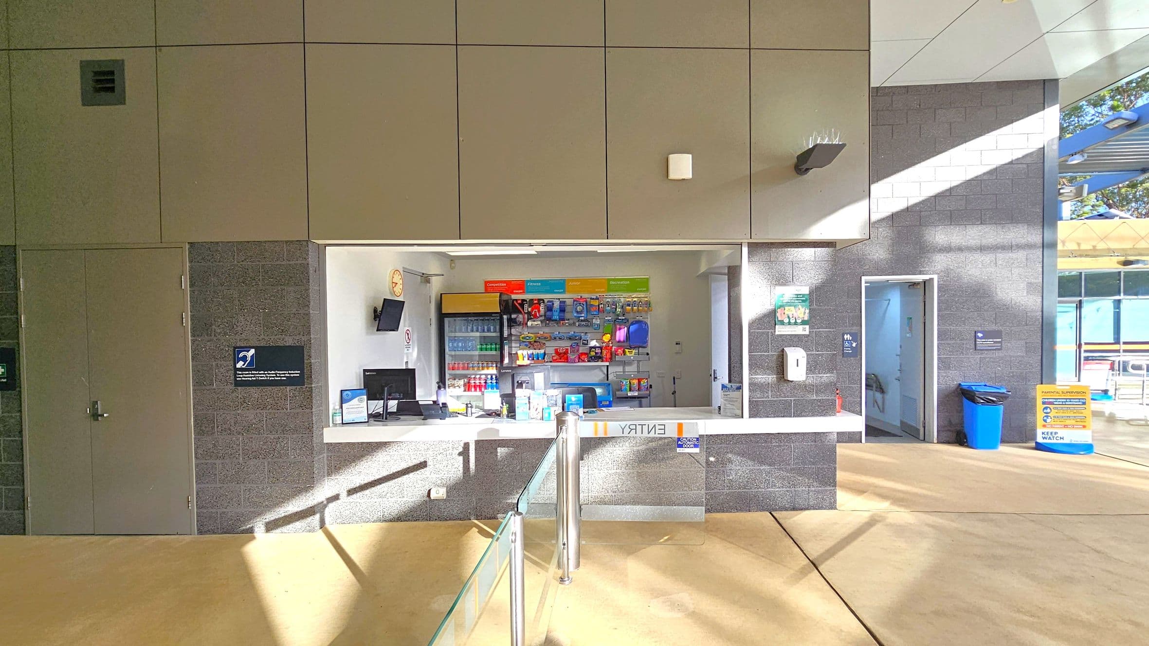 A reception area with a counter in the centre, displaying various items like drinks and snacks on shelves behind. A computer and monitor are on the counter. To the left, a closed door with a sign, and to the right, an open door leading to another room. A blue bin and a yellow sign are near the open door. The floor is light-coloured, and the ceiling has a modern design with panels. Sunlight casts shadows across the scene.