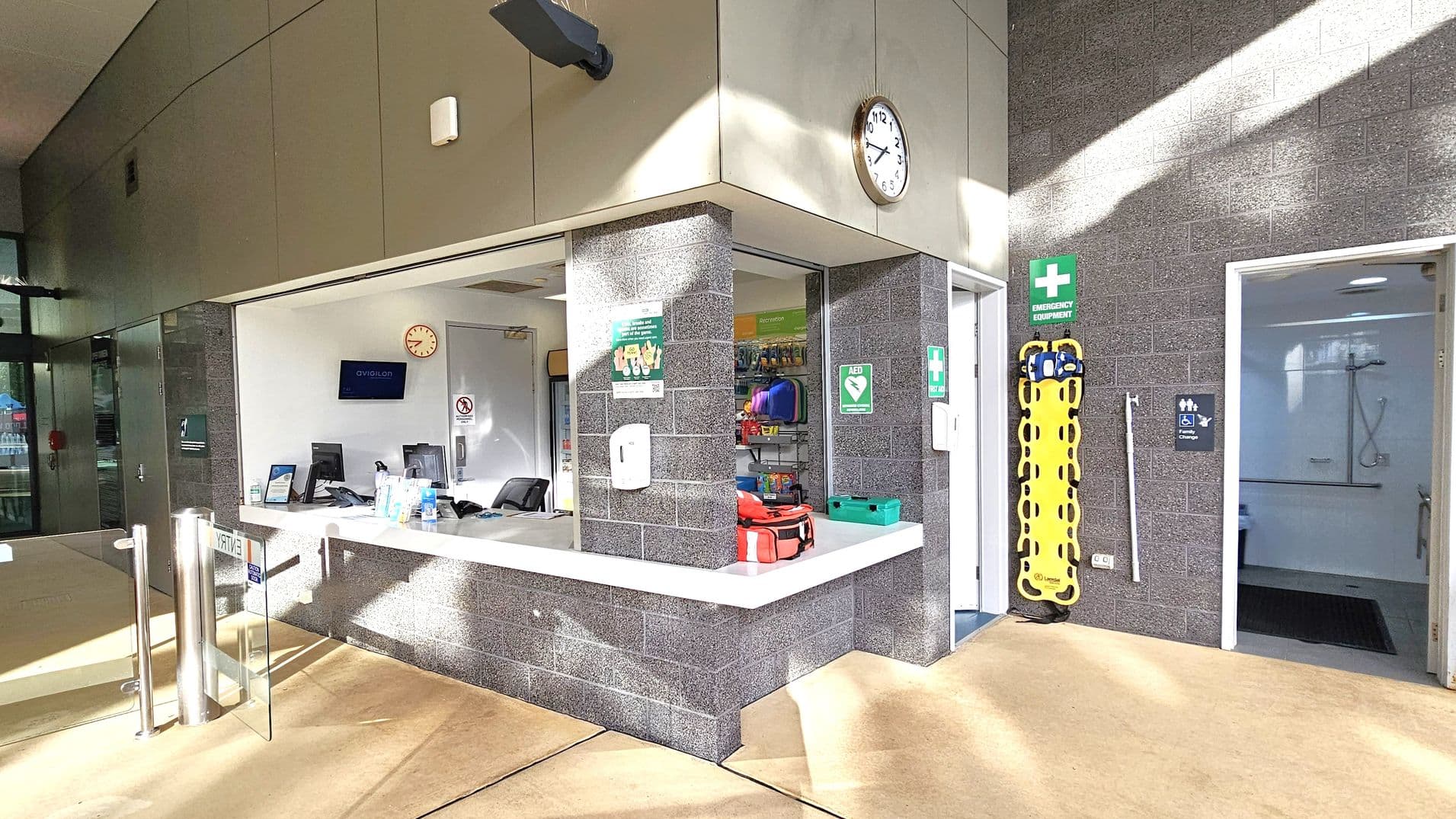 A reception area with a counter in the centre, featuring a computer, hand sanitiser, and informational signs. Behind the counter, a wall clock and a monitor are visible. To the right, a doorway leads to a room with a shower, indicated by a sign. Emergency equipment, including a yellow stretcher, is mounted on the wall. The floor is light-coloured, and the walls are made of grey brick. Sunlight casts shadows across the scene.