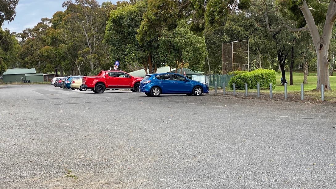 A wide parking space surrounded by trees and marked by short bollards overlooks the entrance to the golf course and the club building to the left. Green-painted metal fence and a golf course fence are seen. At the left end are two green structures.