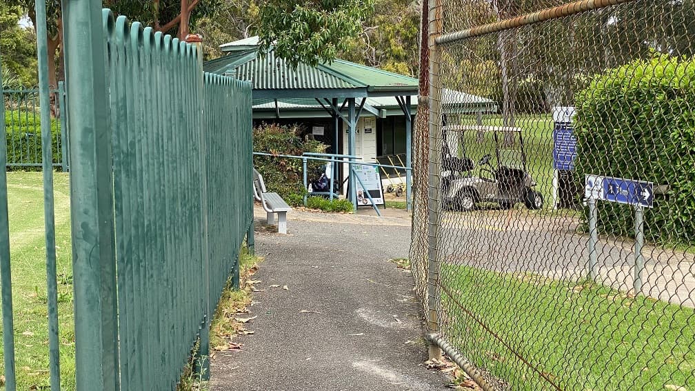 A pathway leads to the golf course features a green-painted metal fence on the left with a banner towards the clubhouse by the end. On the right, a neatly manicured grass fence guides to an entrance marked by short metal bollards. Two direction boards can be seen, along with a golf course fence overlooking the golf course and a red flag pole in the field. The surroundings are adorned with trees and grasses.