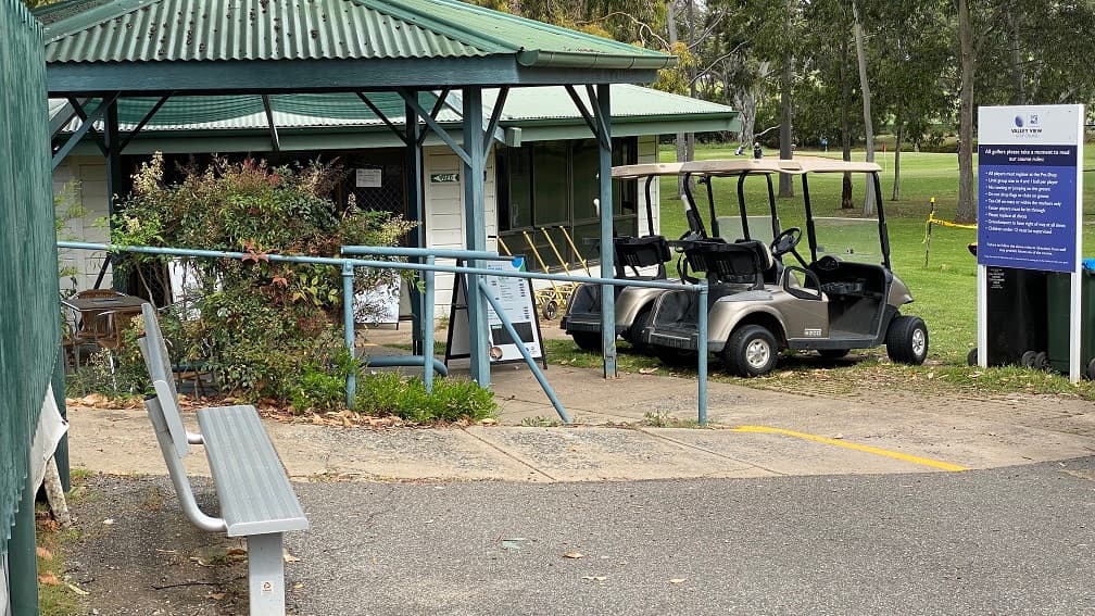 Paved areas located outside the clubhouse with a noticeboard providing directions. On the left, is a long, grey, metal seat by the green-painted metal fences and neatly manicured grass to the right. Two grey golf carts are parked to the right and are facing the green, neatly manicured golf course.