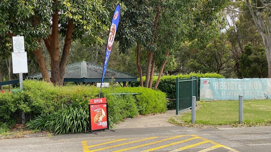 A yellow pedestrian crossing walkway links a footpath by the right with a green-painted metal fence, marketing materials hang from the fence to the right banner and metal bollards mark sections along the path to the right. On the left, the grass conceals the metal fence with signage whilst a sign and banner advertise ice creams. A green roofed structure is further afield.