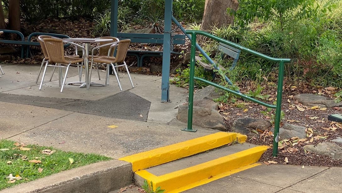 There are two yellow-painted steps with a green rail by the pathway to a sitting area with four brown chairs grouped around a table to the right, overlooking three metal-designed seats covered in dried leaves and grasses. The premise is surrounded by trees and grasslands.