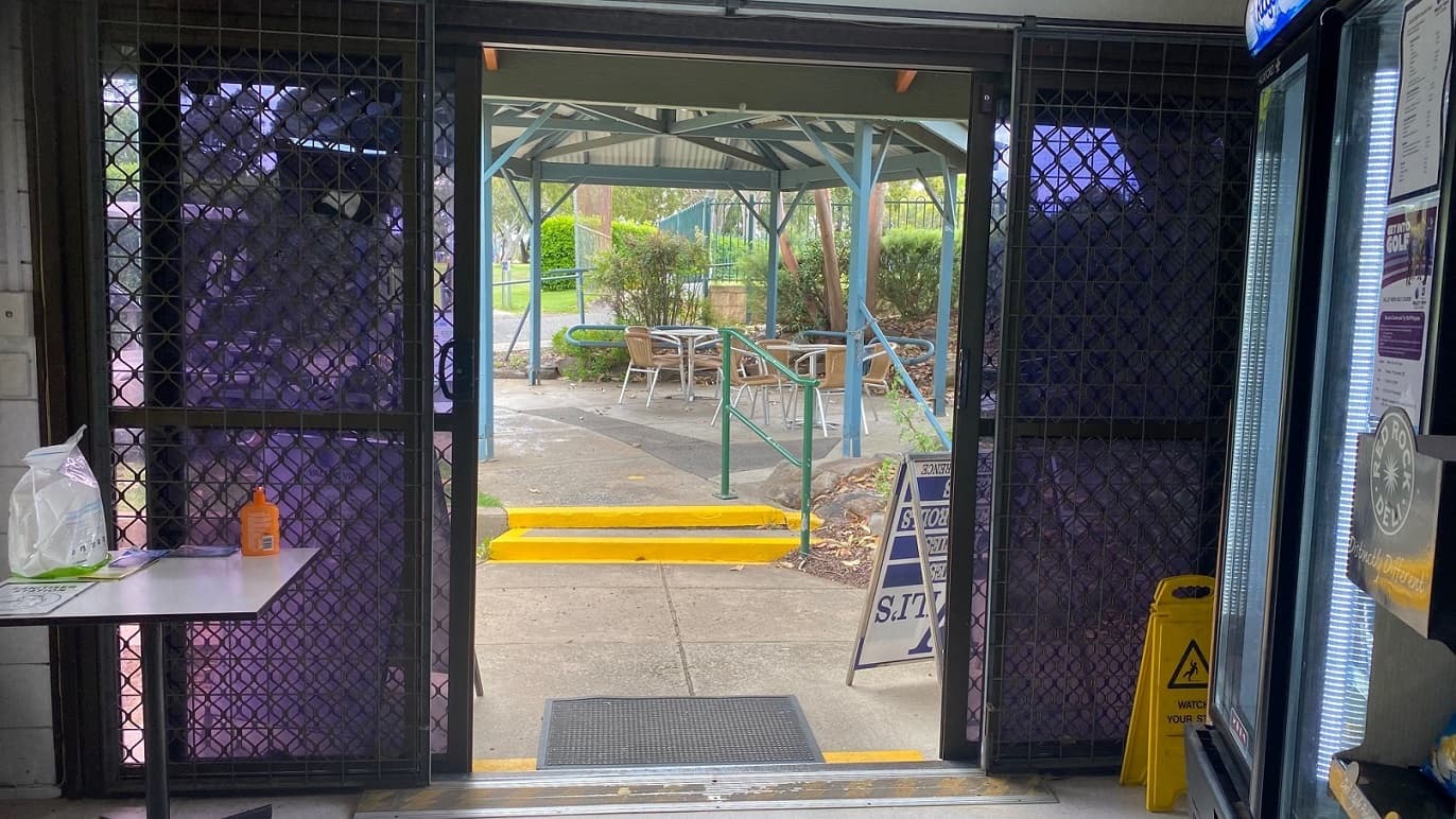 A small white table has brochures, and an orange bottle on it by the left of a dark green sliding door. A drink fridge is to the right and is positioned next to a yellow ‘Watch your step’ sign. Through the open door, there are two yellow steps up to the pavilion with a green balustrade, a sitting area with brown seats, and two tables to the right. At the far end, a green metal fence overlooks a walkway and the golf course. Dried leaves and grassland are in the background.