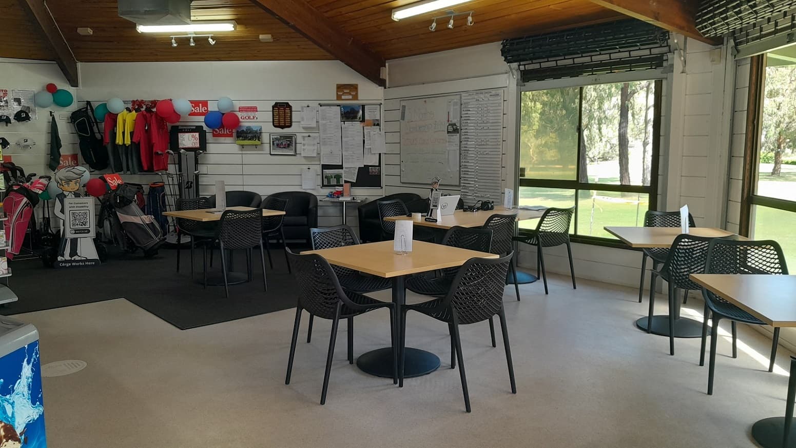 A well-lit tiled retail shop has black and beige flooring. To the right there are black chairs around brown tables, and black sofas, in front of a wall with noticeboard, signage, balloons and retail items for sale. On the black carpet, golf bags, hand carts, club heads & club head covers can be seen with ‘sale’ notices on the wall. On the left, is a freezer next to the checkout desk with a range of snacks for sale. A staff member wearing a black face mask is standing behind the service desk.