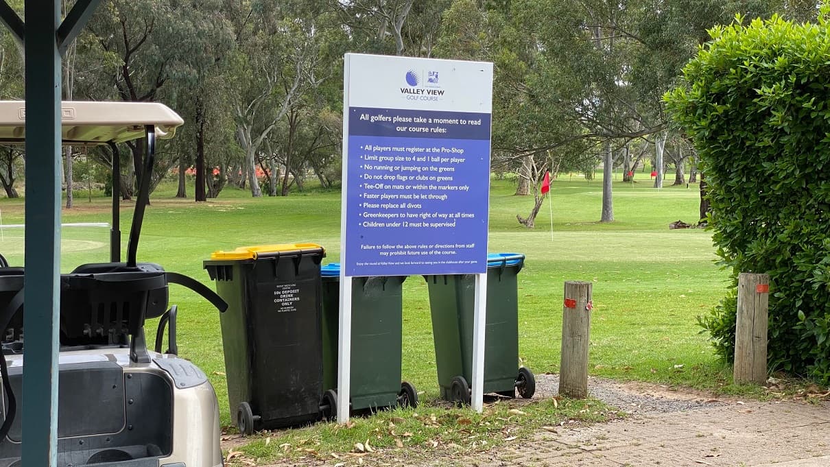 A Valley View golf course notice board stands by two green and a black rubbish bin beside a footpath with wooden bollards and red reflective markers on the right. There is a golf cart on the left. Red flag poles are in the distance. There are trees, flowers, and grasslands all around.