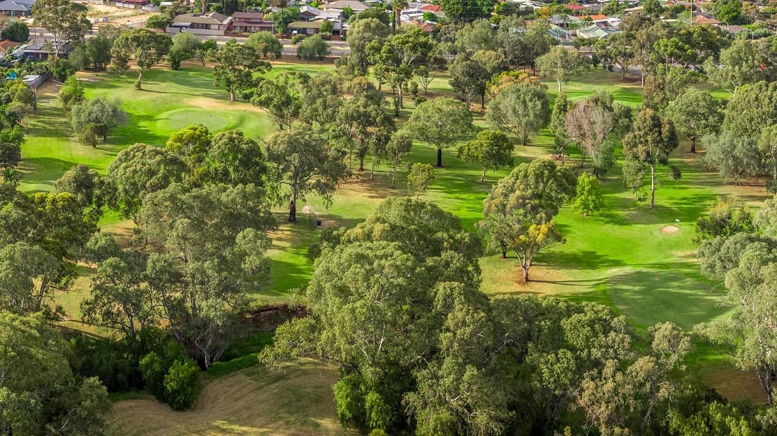 An aerial view of the golf course, with large trees dotted throughout. Towards the back is a residential area with many houses of differing colours and designs.
