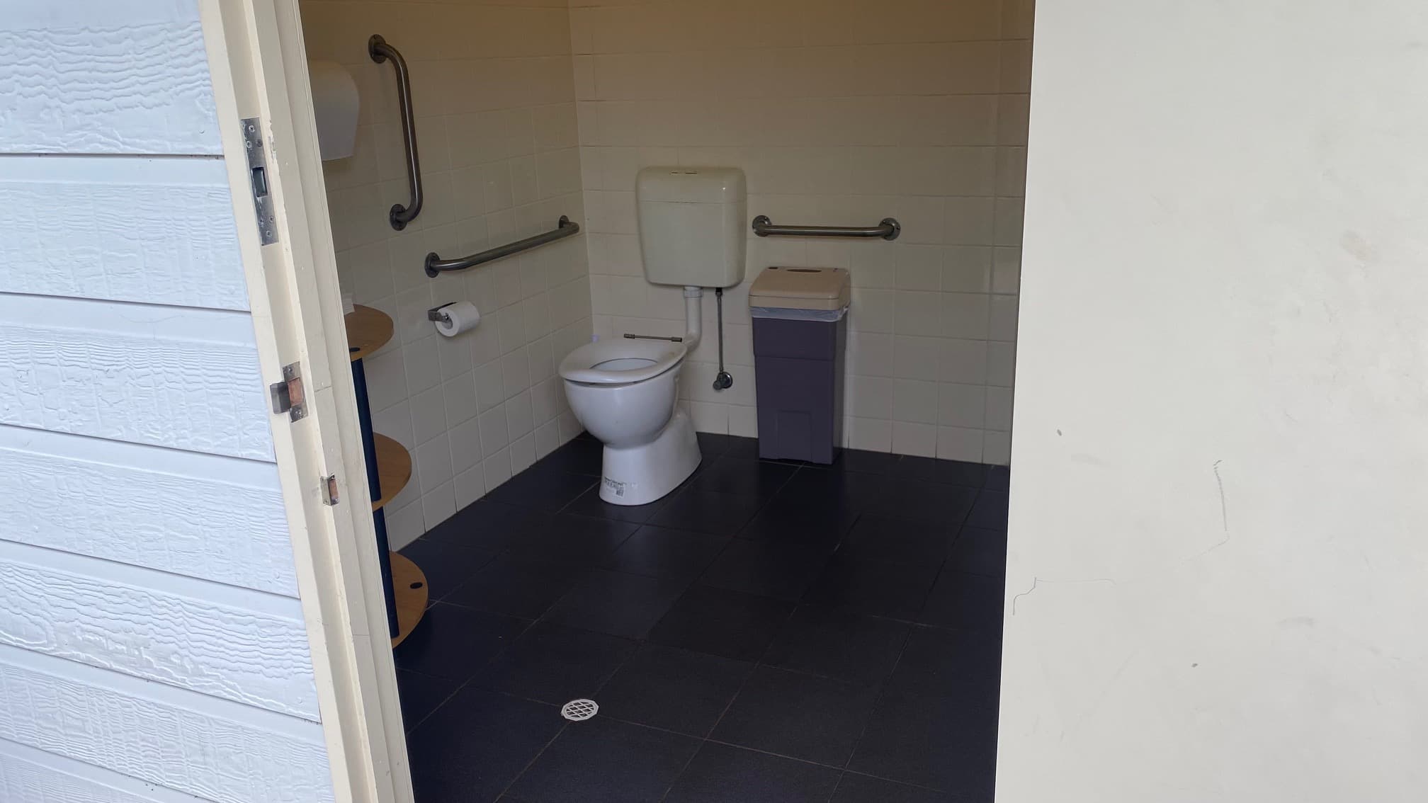 An open door leads to a dark-grey tiled bathroom. There is a sanitary napkin disposal bin to the right of the toilet with three grab rails surrounding the toilet and the toilet roll holder to the left. A paper towel dispenser and a three tiered, brown wooden rack can be seen next to the door.