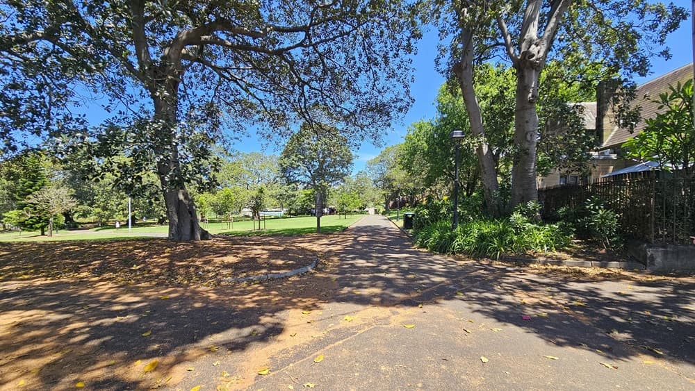 A straight brick path leads forward. To the right, there's a brick house enclosed by a metal fence. The right side is bordered by trees and shrubs. On the left, there's a view of green grass and tall trees.