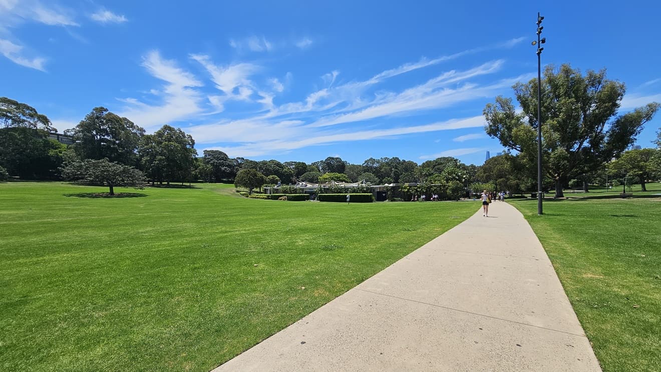 A straight concrete path is bordered by well-maintained grass and tall trees. A tall light post stands on the right. Beyond, there's a facility building. A person walking on the pathway is visible.