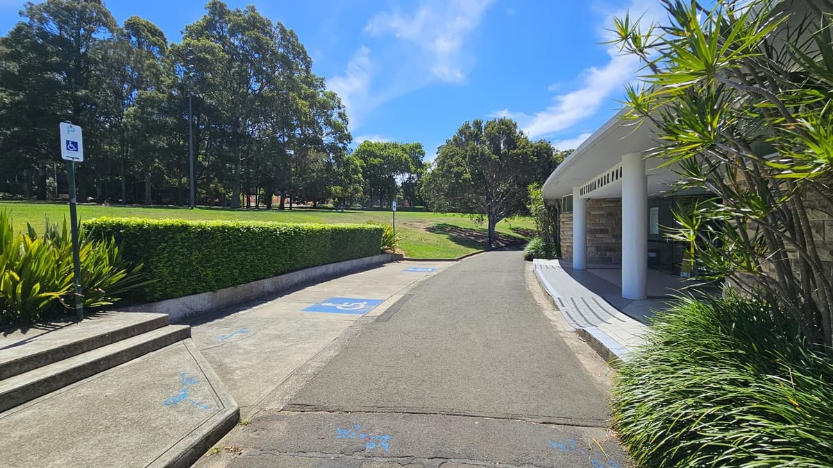 Accessible car parking spaces, marked by blue wheelchair signs, are located in front of the building. To the right, the modern brick structure is equipped with a step-free ramp. In front, there's a landscaped garden and neatly trimmed grass. An accessible sign is positioned on a raised platform near the landscaped shrubs. Tall trees and well-maintained grass surround the space.