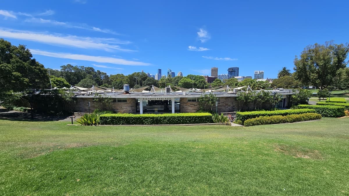 The building is a low-rise modern brick structure with multiple shade cloths at the back. Surrounding the building are landscape shrubs, ornamental plants, and neatly trimmed grass. Further ahead, tall trees and commercial buildings come into view.