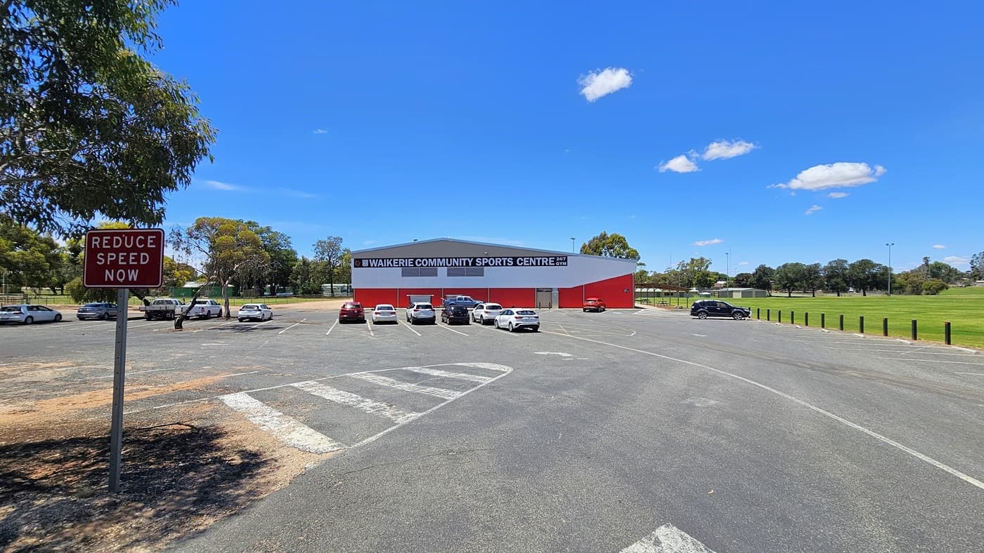 A structure with a red corrugated iron and brown brick exterior stands in the center. Prominent facility signage in bold white letters is displayed. The car park directly adjoins the building, hosting a variety of cars in different models and colours. To the right is a stretch of green grass bordered by concrete bollards. On the left, a road sign indicates "Reduce Speed Now,". The concrete road has white line markings.