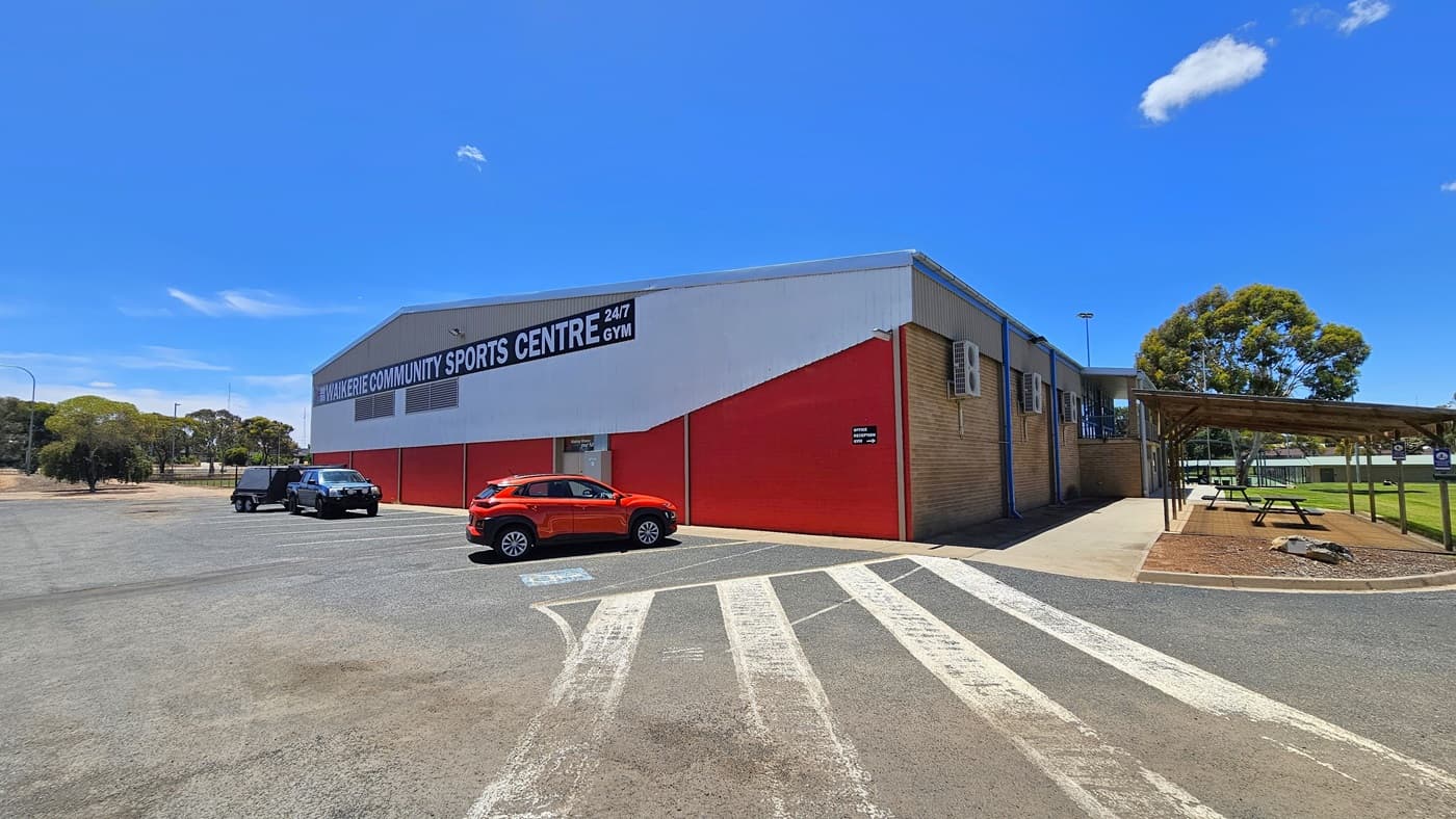 A structure with a red corrugated iron and brown brick exterior stands centrally. Prominent facility signage in bold white letters is displayed. The adjacent concrete car park accommodates three cars of various models and colours, with designated accessible parking on the right, marked by blue wheelchairs and white diagonal lines. To the right of the building is a shaded area with picnic tables, and beyond lies an expanse of green grass. A substantial tree is visible further in.