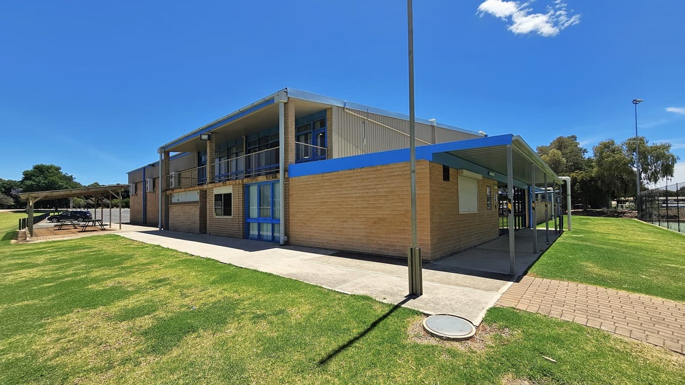 A building with a red corrugated iron and brown brick facade stands centrally. To the left, a shaded area features picnic tables. On the right, a shaded pathway leads to the entrance, and a brick path veers right between the grass. Tall light posts are visible, and a black chain fence surrounds the tennis court.