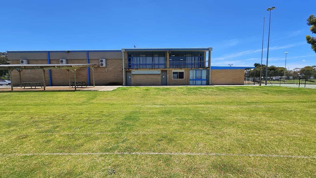 A two-story building with a red corrugated iron and brown brick facade stands in the center. The right wall is made of glass, and condenser units are affixed to the left wall. A shaded area on the left houses two picnic tables. On the right, tall light posts are present, surrounded by green grass in the front, and a tennis court is located on the right.