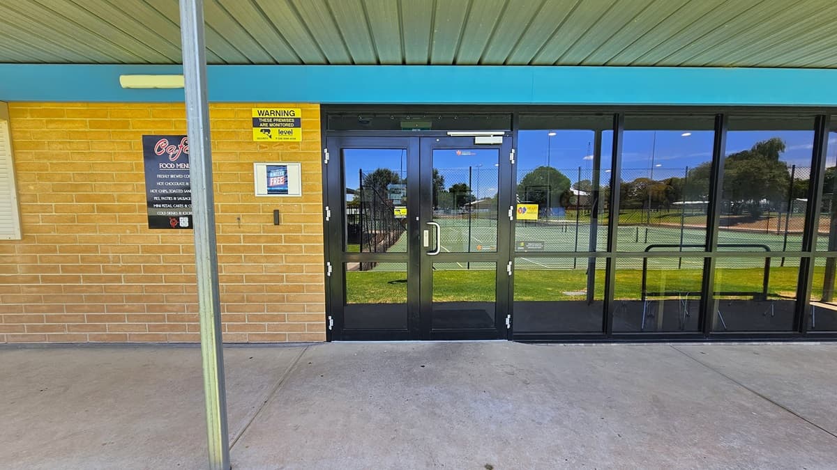 The entrance provides step-free access through a manual glass door opening outward. The right wall is entirely glass, while the left wall is made of brown brick. Informational and safety warning banners are affixed to the left wall. The ceiling features a corrugated design.