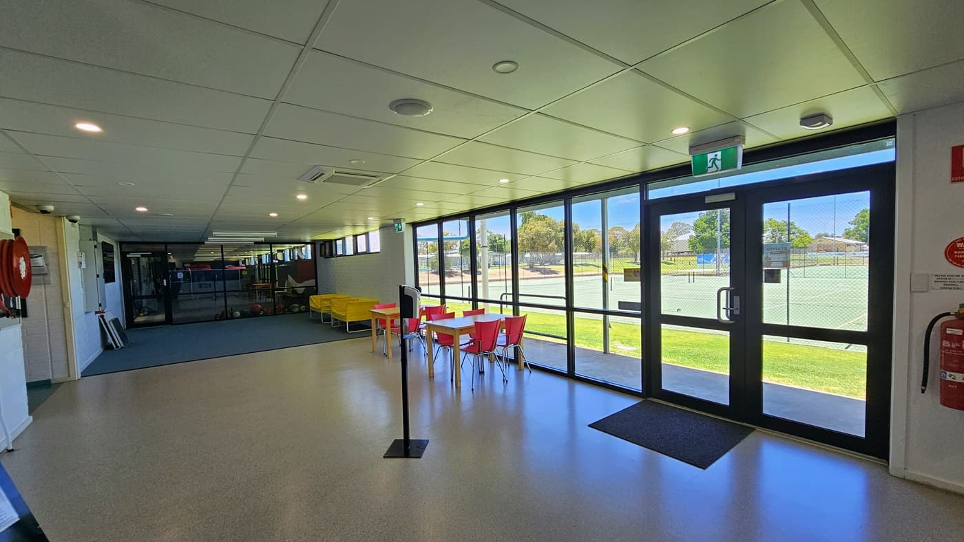 To the right is the glass door equipped with a black anti-slippery mat. Two fire extinguishers are affixed to the wall along with a green exit sign. Movable plastic tables and chairs in yellow and red are positioned on the right near the glass wall. A section of the tennis court outside is visible. On the left is a fire hose reel mounted to the wall. The low ceiling is white with LED lights, and the floor has grey tiles with a granite design.