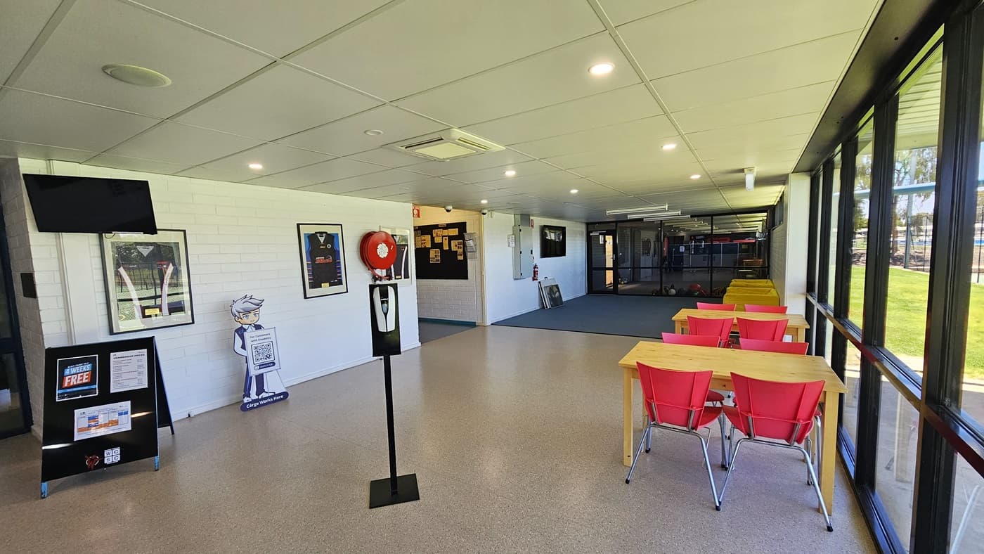 To the right is a floor-to-ceiling glass window allowing a view of the outside. Movable plastic chairs and wooden tables are arranged near the window. On the left is a fire hose reel mounted to the wall along with picture frames. A flat-screen TV is mounted on the wall. A free-standing signage stood underneath. A sanitizer dispenser is placed in the middle. The low ceiling is white with LED lights, and the floor has grey tiles with a granite design.